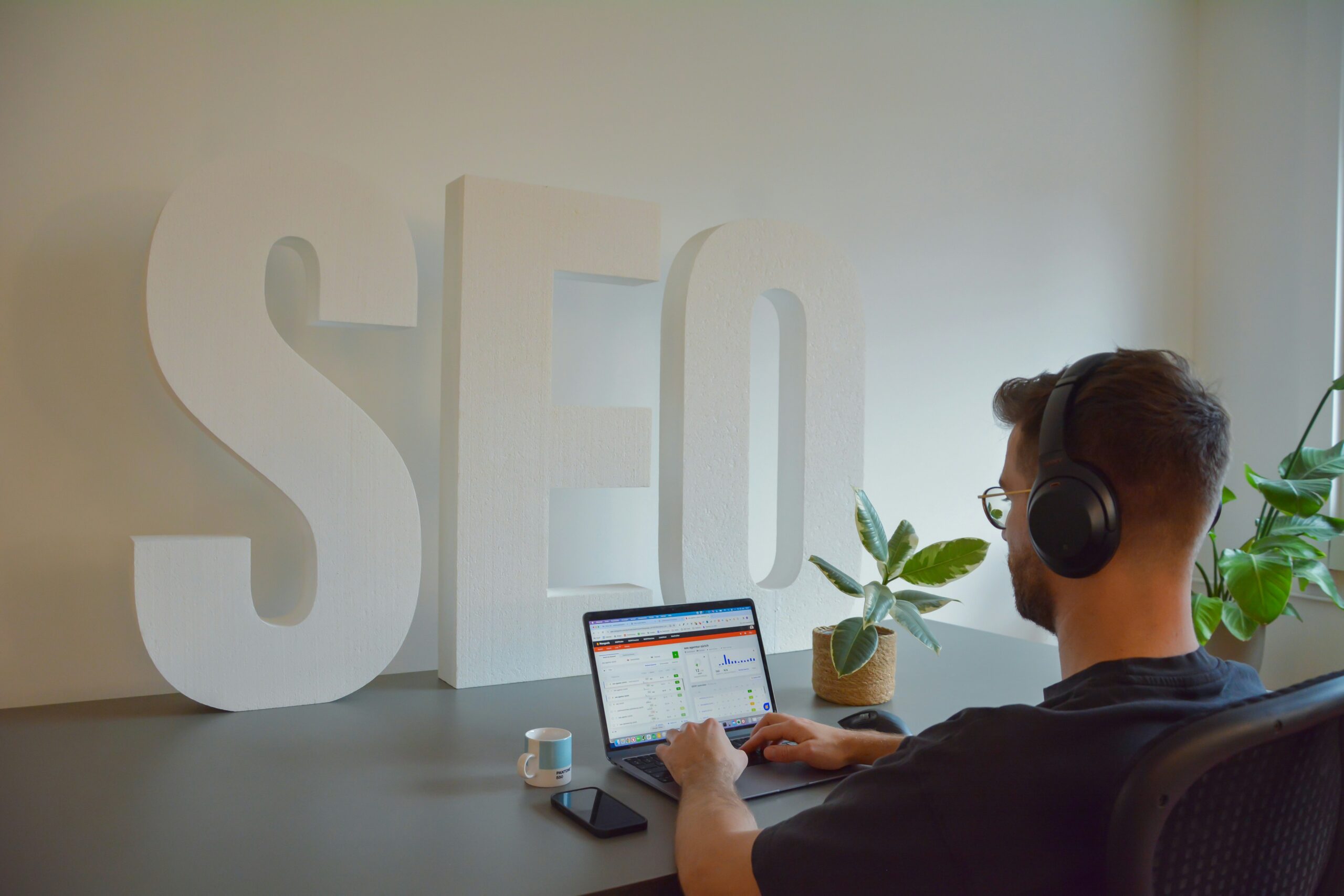 SEO Workspace: A man working on a laptop at a desk with large 3D "SEO" letters and a potted plant in the background.